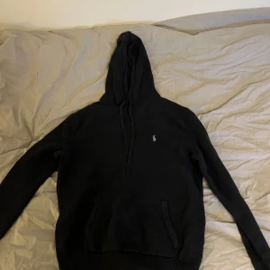 Stickad mörkblå hoodie från Polo Ralph Lauren - Säljer en stilren svart hoodie från Polo Ralph Lauren med det klassiska broderade logotypen på bröstet. Tröjan har en bekväm passform med huva och känguruficka framtill. Perfekt för en avslappnad look.