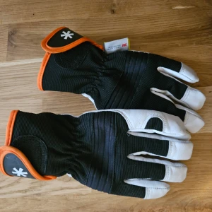 Svart och vit handske från Drytuff - Säljer ett par svarta och vita handskar från Drytuff med orange detaljer. Handskarna har en robust design med förstärkta partier och är isolerade med 3M Thinsulate för extra värme. Perfekta för kalla dagar och utomhusaktiviteter.