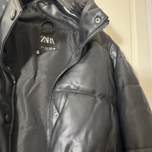 Svart pufferjacka från Zara - Snygg svart pufferjacka från Zara i syntetmaterial. Jackan har en croppad design med dragkedja och knappar framtill. Perfekt för kyligare dagar med sin quiltade stil och elastiska nederkant.