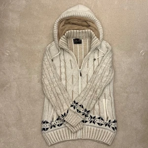 Cardigan med päls  - Säljer en mysig vit stickad cardigan  med dragkedja och ett snyggt svart mönster längst ner. Tröjan har en fodrad huva och ribbade muddar vid ärmslut och nederkant. Perfekt för kyliga dagar! Vid minsta fråga eller fundering hör av er🤝