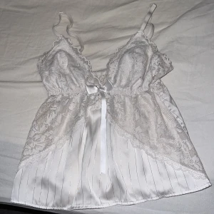 Vit spetslinne med satin och rosett - Elegant vit linne med spetsdetaljer och satinmaterial. Den har smala axelband och en söt rosett framtill. Perfekt för en romantisk kväll eller som en del av en lyxig outfit.