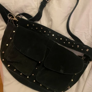 Svart axelväska från Unlimit - Snygg svart axelväska från Unlimit med nitar och flera praktiska fickor. Väskan har en justerbar axelrem och stängs med dragkedja. Perfekt för att ge din outfit en edgy touch.