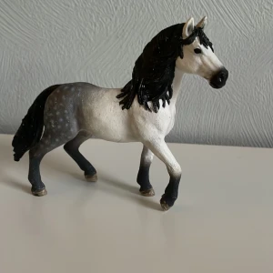 Schleich häst - Detaljerad hästfigur i vit och grå färg med svart man och svans. Figuren har en realistisk design med en stående pose och små prickar på den grå delen av kroppen. Perfekt för samlare eller som dekoration. 