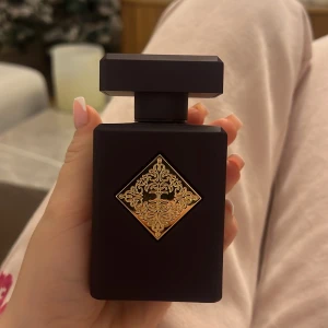 Initio Atomic Rose Parfym - Elegant parfymflaska från Initio med namnet Atomic Rose. Flaskan är mörklila med en dekorativ guldfärgad detalj på framsidan. Noter av italiensk bergamott, bulgarisk ros, turkisk ros, egyptisk jasmin och vanilj från Madagaskar.