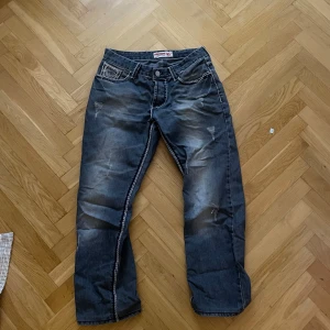 Mörkblå jeans från True Religion - Snygga mörkblå jeans från True Religion med slitna detaljer och kontrastsömmar. Klassisk femficksdesign och knappgylf. Perfekta för en avslappnad stil. Använd ett fåtal gånger men är i väldigt bra skick. Inga skavanker eller nått sånt. W32 L34