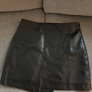 Svart skinnkjol - Snygg svart skinnkjol med omlott-design. Perfekt för att ge en edgy touch till din outfit. Kjolen har en kort längd och en stilren look.