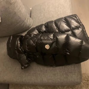 Svart dunjacka från Moncler - Säljer en svart dunjacka från Moncler. Jackan är lång och har en pufferdesign med en huva. Den är utrustad med dragkedja och har Monclers logotyp på arm fickan.HELT OANVÄND. Säljer pga att den e för stor för mig. Perfekt för kalla dagar! ( pris kan diskuteras)
