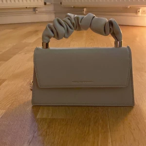Beige handväska med rynkat handtag - Snygg beige handväska med ett unikt rynkat handtag och en elegant kedja för axelrem. Perfekt för att ge en stilren touch till din outfit. Väskan har en enkel och stilren design med en praktisk storlek.