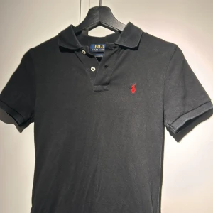 Svart pikétröja från Ralph Lauren - Säljer en stilren svart pikétröja från Ralph Lauren med röd logga på bröstet. Tröjan har korta ärmar och en klassisk krage med knappar. Perfekt för en avslappnad men ändå snygg look.