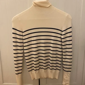 Randig polotröja i beige och svart - Snygg och stilren randig polotröja i beige med svarta ränder. Tröjan har långa ärmar och dekorativa knappar vid ärmsluten. Perfekt för en klassisk look.