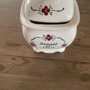 godis skål - Charmig vit keramikburk med lock, dekorerad med röda blommor och blå detaljer. Texten 'Smått & Gott' pryder framsidan. Perfekt för att förvara småsaker eller som en söt inredningsdetalj i köket.
