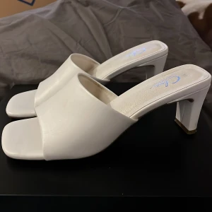 Vita klackskor - Snygga vita klackskor i syntet med bred rem över foten. Klacken är 6,5cm hög. Passar bra till både klänning och jeans.