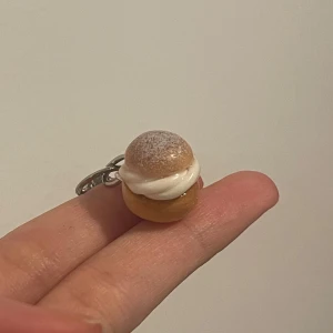 Nyckelring med semla - Supergullig nyckelring formad som en semla! Den har en realistisk look med en liten bulle, vit grädde och pudrat socker på toppen. Perfekt för att piffa upp dina nycklar eller väskan. Perfekt present till någon som älskar bakverk! 🥰