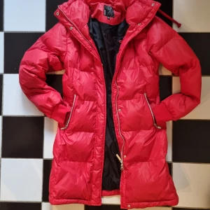 Röd dunjacka från Stadium - Säljer en snygg röd dunjacka från Stadium. Jackan är lång och har en dragkedja framtill samt praktiska fickor med dragkedjor. Perfekt för kalla vinterdagar med sitt varma dunfoder. Yttermaterialet är 100% nylon vilket gör den både lätt och slitstark. Passar perfekt för vinterns alla äventyr! 🔥