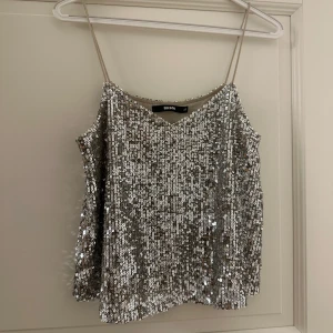 Silverfärgad paljettopp från Bikbok - Säljer en glittrig silverfärgad paljettopp från Bikbok. Den har tunna axelband och en v-ringad design, perfekt för festliga tillfällen. Toppen är i mycket bra skick och ger en glamorös touch till vilken outfit som helst. Perfekt för nyår eller en utekväll! ✨