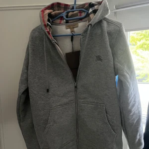 Burberry zip hoddie - Säljer en snygg grå huvtröja från Burberry i helt ny skick prislapp kvar!!. Den har en dragkedja framtill och en klassisk rutig insida i huvan. Perfekt för höst och vår, med en normal passform och långa ärmar. Superbekväm och stilren!