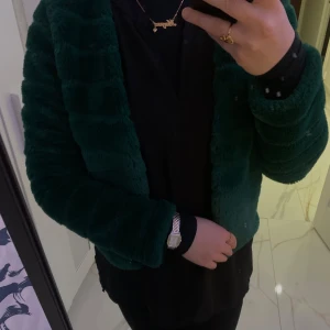 Mörkgrön pälsjacka - Säljer en supermjuk och mysig mörkgrön pälsjacka. Den har en elegant design med långa ärmar och en enkel stängning framtill. Perfekt för att hålla sig varm och stilren under kyliga dagar. Passar både till vardags och festliga tillfällen.