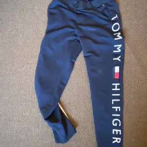 Säljer ett par snygga blå mjukisbyxor från Tommy Hilfiger. De har en bekväm passform med resår i midjan och muddar vid bensluten. Stort Tommy Hilfiger-tryck längs benet ger en cool look. Perfekta för en avslappnad dag hemma eller en snabb tur till affären.