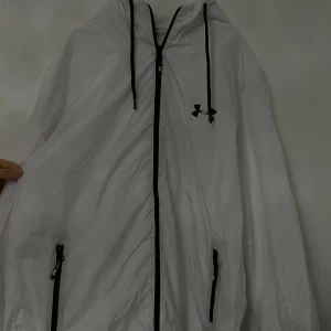 Vit vindjacka från Under Armour - Säljer en vit vindjacka från Under Armour i mycket bra skick. Jackan har en dragkedja framtill och justerbar huva med svarta snören. Perfekt för blåsiga dagar och sportiga aktiviteter. Den är lätt och bekväm att bära. Passar S/M