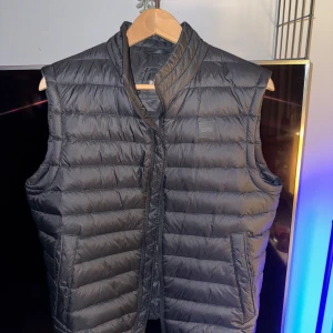 Svart dunväst från Boss - Säljer en snygg svart dunväst från Boss i storlek M. Västen är quiltad och har en dragkedja framtill. Perfekt för höst och vår när du vill hålla dig varm men ändå se stilren ut. Den har praktiska fickor med knappar för extra säkerhet. Passar både till vardags och fest!