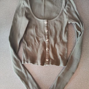 Olivgrön ribbad topp från Gina Tricot - Säljer en superfin olivgrön ribbad topp från Gina Tricot. Den har långa ärmar och knappar framtill, vilket ger en snygg look. Perfekt för både vardag och fest!