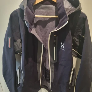Svart och grå vindjacka från Haglöfs - Säljer en svart och grå vindjacka från Haglöfs. Jackan har en praktisk huva och flera dragkedjeförsedda fickor. Den är utrustad med en RECCO Avalanche Rescue Reflector, vilket gör den perfekt för äventyr i bergen. Jackan är i bra skick och redo för nya äventyr!