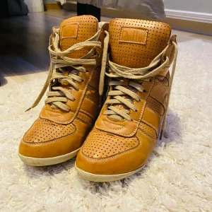 Bruna sneakers med kilklack från Duffy - Säljer ett par snygga bruna sneakers från Duffy i mycket bra skick. Stl 38!De har en cool kilklack och perforerat mönster för extra stil. Skorna är i skinn och har snörning framtill. Perfekta för höst och vår när du vill ha både komfort och stil.