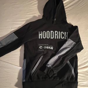Hoodrich hoodie - Säljer en snygg svart och grå hoodie från Hoodrich med tryck både fram och bak. Den har en stor ficka framtill och en bekväm huva. Perfekt för en casual look eller streetwear-stil. Passar bra till både jeans och joggers!