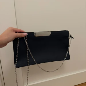 Svart kuvertväska med kedja - Snygg svart kuvertväska i syntetmaterial med en elegant kedja som axelrem. Väskan har en stilren design med en dragkedja upptill och en metallisk detalj på framsidan. Perfekt för festliga tillfällen eller en utekväll. HELT OANVÄND