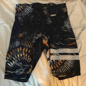 Svarta träningsshorts från Stronger - Säljer ett par coola svarta träningsshorts från Stronger med ett unikt mönster i orange, vitt och grått. De har en bekväm passform och är perfekta för gymmet eller löprundan. Shortsen har en elastisk midja och är gjorda i ett stretchigt material för bästa komfort. Strl L