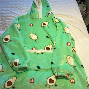 Grön fleece hoodie med avokadomönster - Supermysig och rolig grön hoodie i fleece med gulligt avokadomönster. Perfekt för kalla dagar när du vill vara både varm och stilfull. Den har en stor ficka fram och en fluffig insida som känns som att ha på sig en kram. Perfekt för hemmamys eller som en rolig present!