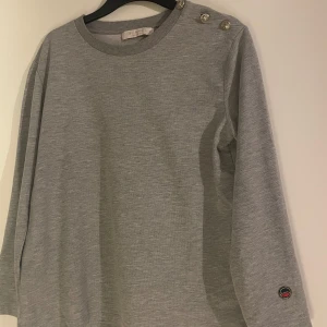 Grå tröja från Busnel - Säljer en stilren grå tröja från Busnel i mycket bra skick. Tröjan har långa ärmar och dekorativa knappar på axeln. Perfekt för både vardag och lite finare tillfällen. Den är mjuk och bekväm, vilket gör den idealisk för höst och vår.