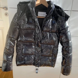 Svart pufferjacka från Calvin Klein Jeans - Säljer en snygg svart pufferjacka från Calvin Klein Jeans i storlek XS. Jackan har en glansig finish och är perfekt för kalla dagar. Den har en dragkedja framtill och en mysig luva. Perfekt för både vardag och festliga tillfällen!