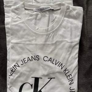 Säljer en snygg vit t-shirt från Calvin Klein Jeans. Den har en klassisk rund hals och ett stort tryck med 'CK' och 'Calvin Klein Jeans' i svart på framsidan. Perfekt för en casual look eller att ha under en jacka. Passar till alla tillfällen!