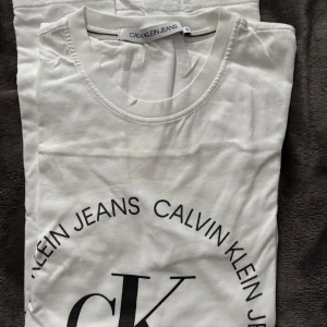 Vit t-shirt från Calvin Klein Jeans - Säljer en snygg vit t-shirt från Calvin Klein Jeans. Den har en klassisk rund hals och ett stort tryck med 'CK' och 'Calvin Klein Jeans' i svart på framsidan. Perfekt för en casual look eller att ha under en jacka. Passar till alla tillfällen!