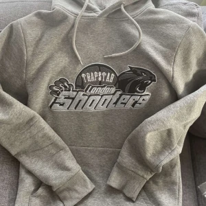 Grå hoodie från Trapstar - Säljer en grå hoodie från Trapstar med trycket 'London Shooters' och en cool panterdesign på framsidan. Den har en klassisk känguruficka och justerbar huva med snören. Perfekt för en avslappnad stil eller streetwear-look. Superbekväm och i bra skick!