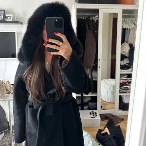 Svart kappa med pälshuva - Säljer en elegant svart kappa med en lyxig pälshuva. Den har en knytning i midjan för en snygg passform och är perfekt för kalla vinterdagar. Jackan är långärmad och i mycket bra skick. Perfekt för både vardag och festliga tillfällen!