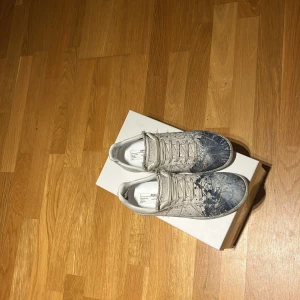 Maison Margiela sneakers med blått mönster - Säljer ett riktigt eftertraktade Maison Margiela sneakers i mycket bra skick. Skorna är vita med ett coolt blått mönster som ger dem en konstnärlig touch. De har klassisk snörning och kommer i originalförpackning. Perfekta för dig som vill sticka ut med stil!