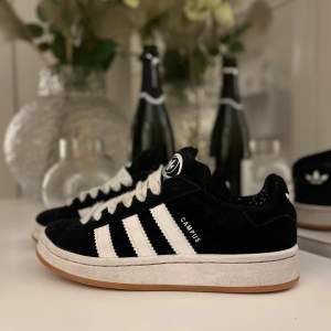 Säljer ett par svarta Adidas Campus sneakers i mocka med vita detaljer och snörning. De har en klassisk design med tre vita ränder på sidan och en vit sula. Skorna är i bra skick men har lite slitage på insidan. Perfekta för både vardag och fest!