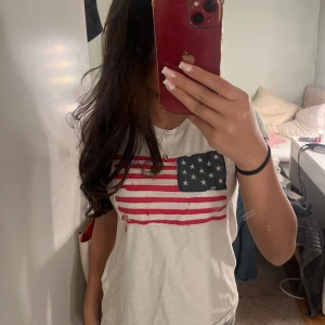 Vit t-shirt med amerikansk flagga - Säljer en vit t-shirt från Ralph Lauren med en cool amerikansk flagga på bröstet. Perfekt för en avslappnad stil. T-shirten är kortärmad och i bomull, vilket gör den superbekväm att ha på sig. Passar perfekt till jeans eller shorts för en casual look.