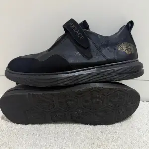 Snygga svarta sneakers från Versace i mycket bra skick. De har en stilren design med kardborreband och en diskret logga på sidan. Perfekta för både vardag och festliga tillfällen. Skorna är gjorda i skinn och mocka vilket ger en lyxig känsla.