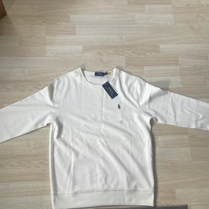 Vit tröja från Polo Ralph Lauren - Säljer en stilren vit tröja från Polo Ralph Lauren i nytt skick. Tröjan har en klassisk design med långärmade ärmar och det ikoniska logotypbroderiet på bröstet. Perfekt för både vardag och festliga tillfällen. Passar bra till jeans eller chinos för en avslappnad men ändå elegant look.