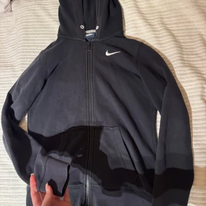 Svart hoodie från Nike - Säljer en svart hoodie från Nike i mycket bra skick. Den har en dragkedja framtill och en klassisk Nike-logga på bröstet. Perfekt för höst och vår, och den är superbekväm med sin mjuka bomullsblandning. Passar både till träning och vardagsbruk.