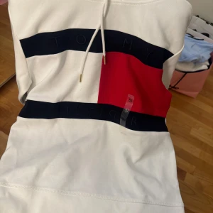 Vit hoodie från Tommy Hilfiger - Säljer en snygg vit hoodie från Tommy Hilfiger med deras ikoniska logga i rött, vitt och blått på framsidan. Den har en huva med snören och är i nytt skick. 