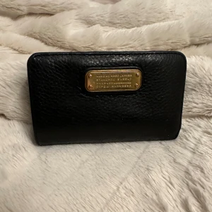 Svart plånbok från Marc Jacobs - Säljer en elegant svart plånbok från Marc Jacobs i skinn. Den har flera kortfack och en snygg guldfärgad logotyp på framsidan. Perfekt för att hålla ordning på kort och kontanter med stil. Plånboken är i  bra skick, dock lite skavd på insidan - se bild 2, och har en klassisk design som passar alla tillfällen. Inköpt 2019. 