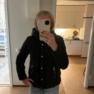 Svart vinterjacka - Varm, bekväm, svart vinterjacka med guldiga detaljer. Rimlig luva och medellängd. Använd fåtal gånger. Två fickor med dragkedja.