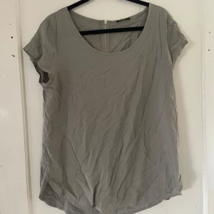 Grå topp från Colloseum - Säljer en enkel och stilren grå topp från Colloseum i storlek XL. Toppen har korta ärmar och en rund halsringning. Den är gjord i ett mjukt material och har en dragkedja i ryggen för en snygg detalj. Perfekt för vardagsbruk!