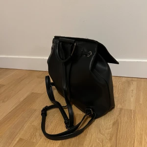Svart liten ryggsäck  - Svart liten ryggsäck i läderimitation. Har ett litet innerfack för värdesaker och axelband som går att justera. Endast använd fåtal gånger. 