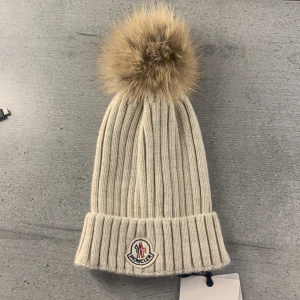 Helt ny beige ribbad mössa från Moncler - Snygg beige mössa från Moncler med ribbad design och en stor pälsboll på toppen. Mössan har Monclers logotyp framtill och är perfekt för kyliga dagar. Helt ny med lappen fortfarande på