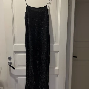 Svart glittrig klänning med tunna axelband - Elegant svart klänning med glittrigt tyg och tunna axelband. Perfekt för en festlig kväll. Klänningen har en enkel och stilren design som ger en sofistikerad look.